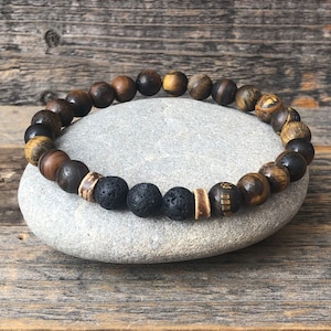 May include: A beaded bracelet featuring a mix of dark brown, tiger's eye, and black lava stone beads. The bracelet is strung on an elastic cord and rests on a gray stone, showcasing the natural variations in the materials.