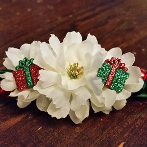 May include: A festive hair accessory featuring white fabric flowers with two glittery gift box embellishments in red and green. The accessory has a green and red ribbon base, perfect for holiday celebrations.