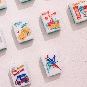 May include: A collection of white rectangular mahjong tiles featuring colorful illustrations and text. Designs include a red Hong Kong skyline, a Dim Sum basket, a blue bird, a red truck with the text Have a Sweet Day, and a red mailbox.