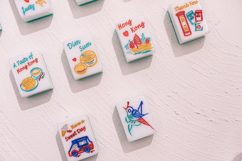 May include: A collection of white rectangular mahjong tiles featuring colorful illustrations and text. Designs include a red Hong Kong skyline, a Dim Sum basket, a blue bird, a red truck with the text Have a Sweet Day, and a red mailbox.