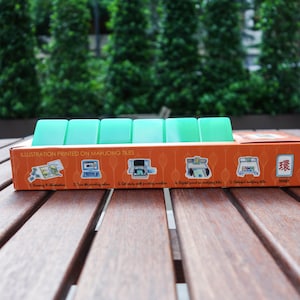 Peut inclure: Une bo&icirc;te de jeu de mahjong avec une rang&eacute;e de tuiles vert clair. La bo&icirc;te est orange avec des illustrations et le texte "ILLUSTRATION PRINTED ON MAHJONG TILES". L'arri&egrave;re-plan est une table en bois et un feuillage vert.