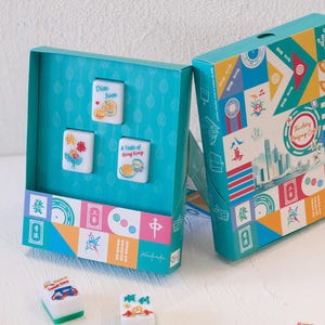 May include: A teal box set featuring three white mahjong tiles with colorful illustrations and text including Dim Sum and A Taste of Hong Kong. The box exterior displays a vibrant geometric pattern with various icons and the text The Art of Mahjong Craft.