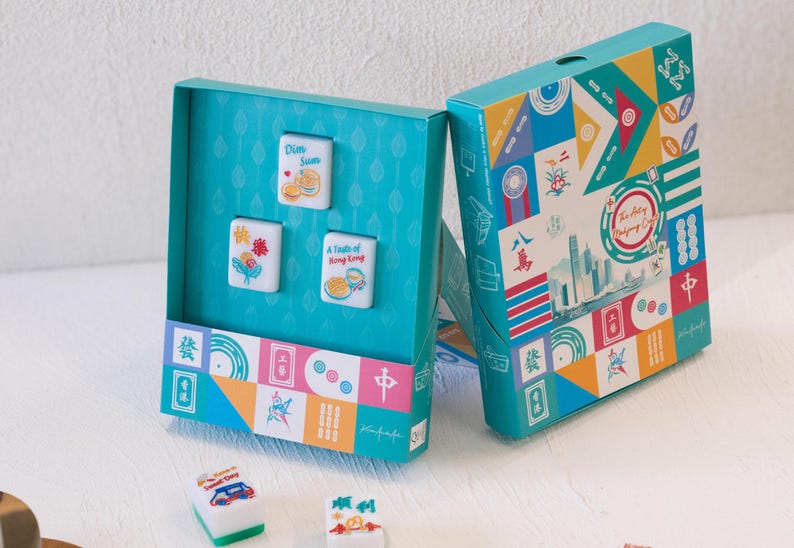 May include: A teal box set featuring three white mahjong tiles with colorful illustrations and text including Dim Sum and A Taste of Hong Kong. The box exterior displays a vibrant geometric pattern with various icons and the text The Art of Mahjong Craft.