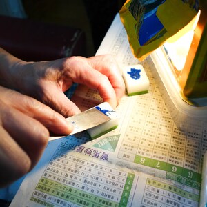 Peut inclure: Une personne utilise une petite lame pour appliquer de l'encre bleue sur une tuile blanche et verte. Plusieurs tuiles sont visibles, ainsi qu'un r&eacute;cipient et un journal avec des caract&egrave;res chinois.