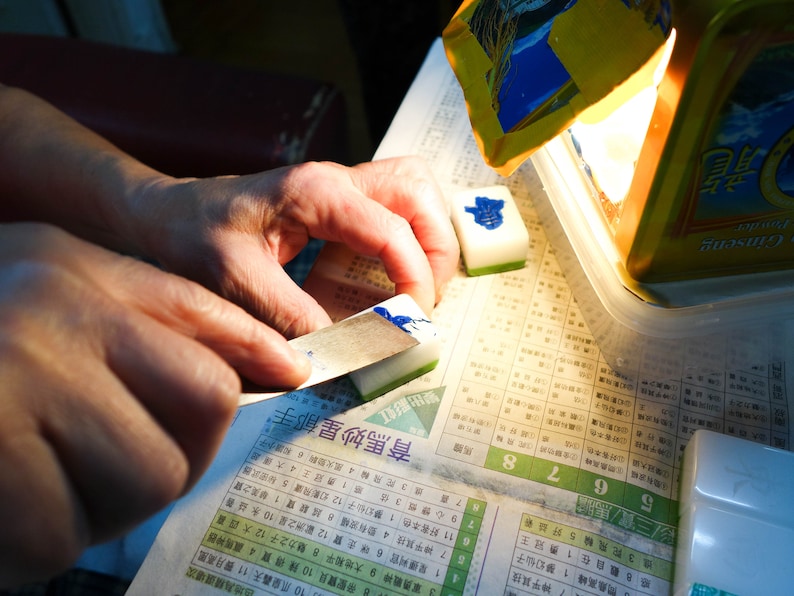 Peut inclure: Une personne utilise une petite lame pour appliquer de l'encre bleue sur une tuile blanche et verte. Plusieurs tuiles sont visibles, ainsi qu'un r&eacute;cipient et un journal avec des caract&egrave;res chinois.