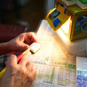 Peut inclure: Une personne utilise un petit couteau avec un manche rouge et jaune pour travailler sur un objet rectangulaire blanc et vert. L'objet est sur un journal avec des caract&egrave;res chinois. Un r&eacute;cipient dor&eacute; avec du texte chinois est en arri&egrave;re-plan.