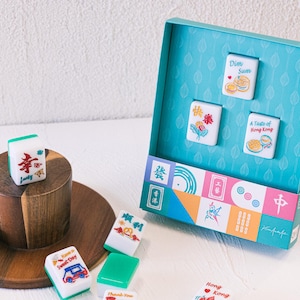 May include: A set of white and green mahjong tiles featuring colorful illustrations and text including Dim Sum, A Taste of Hong Kong, and Happy Life. The tiles are displayed both loose on a wooden stand and inside a teal box with a patterned interior.