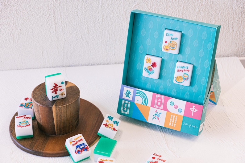 May include: A set of white and green mahjong tiles featuring colorful illustrations and text including Dim Sum, A Taste of Hong Kong, and Happy Life. The tiles are displayed both loose on a wooden stand and inside a teal box with a patterned interior.
