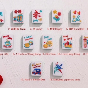 May include: Thirteen white rectangular mahjong tiles arranged in rows, each featuring colorful illustrations and text including phrases like Lucky, Happy, Dim Sum, and Hong Kong. Two paintbrushes and a red bird figurine are visible on the left.