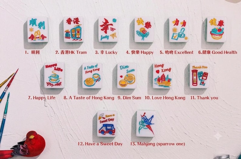 May include: Thirteen white rectangular mahjong tiles arranged in rows, each featuring colorful illustrations and text including phrases like Lucky, Happy, Dim Sum, and Hong Kong. Two paintbrushes and a red bird figurine are visible on the left.