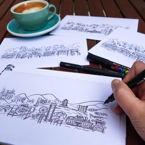 Peut inclure: Une main esquisse un paysage urbain d&eacute;taill&eacute; en noir et blanc sur un carnet &agrave; spirales blanc. Plusieurs autres croquis de paysages urbains sont sur la table, ainsi que des marqueurs noirs et une tasse de caf&eacute; dans une tasse et une soucoupe turquoise.