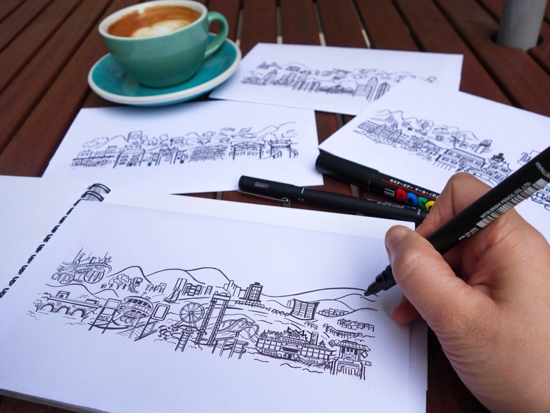 Peut inclure: Une main esquisse un paysage urbain d&eacute;taill&eacute; en noir et blanc sur un carnet &agrave; spirales blanc. Plusieurs autres croquis de paysages urbains sont sur la table, ainsi que des marqueurs noirs et une tasse de caf&eacute; dans une tasse et une soucoupe turquoise.