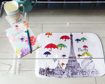 Op de afbeelding: Een witte deurmat met een zwart-witte illustratie van de Eiffeltoren en het woord "Paris". De deurmat is versierd met kleurrijke parapluies.