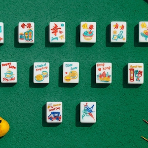 May include: A collection of thirteen rectangular white mahjong tiles arranged in three rows, each featuring colorful illustrations and text including phrases like Happy Life, Dim Sum, Hong Kong, and Thank You, alongside a small yellow bird figurine.