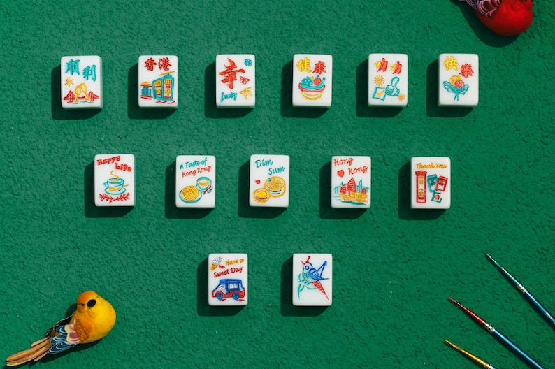 May include: A collection of thirteen rectangular white mahjong tiles arranged in three rows, each featuring colorful illustrations and text including phrases like Happy Life, Dim Sum, Hong Kong, and Thank You, alongside a small yellow bird figurine.
