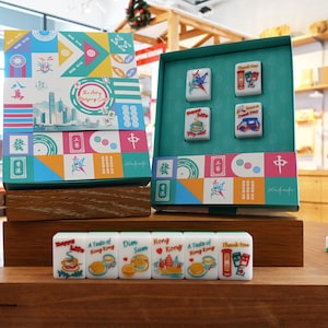 May include: A Mahjong set featuring a colorful box with geometric patterns and a cityscape illustration. The set includes white tiles with various food and Hong Kong-themed graphics, including text like "Happy Life," "Dim Sum," "Hong Kong," and "Thank You."