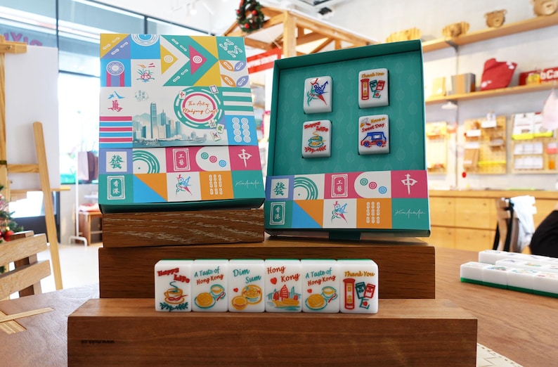 May include: A Mahjong set featuring a colorful box with geometric patterns and a cityscape illustration. The set includes white tiles with various food and Hong Kong-themed graphics, including text like "Happy Life," "Dim Sum," "Hong Kong," and "Thank You."