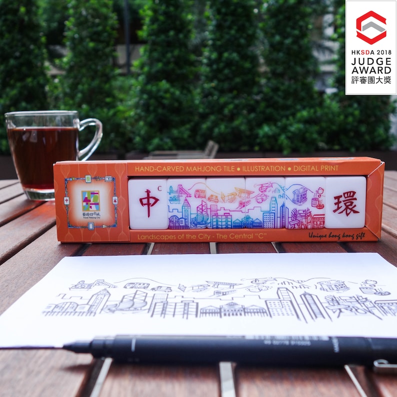 Peut inclure: Un coffret de tuiles de mahjong sculpt&eacute;es &agrave; la main avec une illustration de paysage urbain. Les tuiles blanches pr&eacute;sentent des motifs de skyline color&eacute;s et des caract&egrave;res chinois. La bo&icirc;te orange porte l'inscription "Landscapes of the City - The Central 'C'".