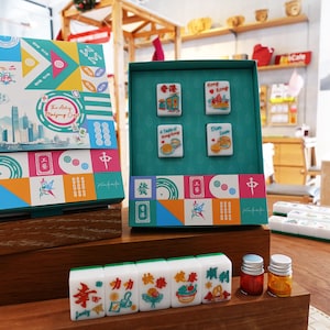 May include: A mahjong set featuring a colorful box with geometric patterns and a skyline illustration labeled The Artsy Mahjong Club. The set includes four tiles with Hong Kong themed graphics and a row of five tiles with Chinese characters and English text.