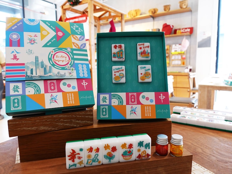 May include: A mahjong set featuring a colorful box with geometric patterns and a skyline illustration labeled The Artsy Mahjong Club. The set includes four tiles with Hong Kong themed graphics and a row of five tiles with Chinese characters and English text.