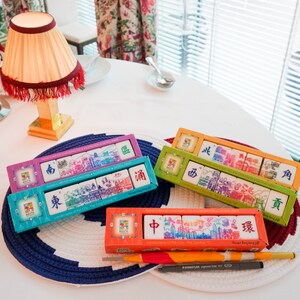 Peut inclure: Quatre bo&icirc;tes de tuiles de Mahjong sculpt&eacute;es &agrave; la main avec des illustrations de paysages urbains. Chaque bo&icirc;te contient des tuiles avec des caract&egrave;res chinois et des sc&egrave;nes de ville. Les bo&icirc;tes sont violettes, vertes, bleues et oranges, sur une table blanche.