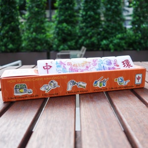 Peut inclure: Un ensemble de tuiles de Mahjong blanches avec des symboles et des illustrations rouges et bleus, pr&eacute;sent&eacute; dans une bo&icirc;te orange. La bo&icirc;te contient des instructions pour sculpter les tuiles &agrave; la main. Les tuiles sont sur une table en bois avec un fond vert.