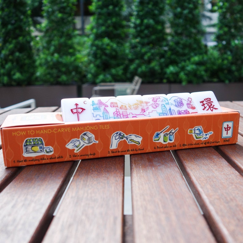 Peut inclure: Un ensemble de tuiles de Mahjong blanches avec des symboles et des illustrations rouges et bleus, pr&eacute;sent&eacute; dans une bo&icirc;te orange. La bo&icirc;te contient des instructions pour sculpter les tuiles &agrave; la main. Les tuiles sont sur une table en bois avec un fond vert.