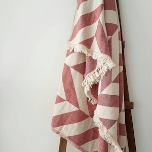 May include: A red and cream geometric patterned throw blanket with fringe trim is draped over a wooden ladder. The blanket features a repeating pattern of angled stripes and triangles.