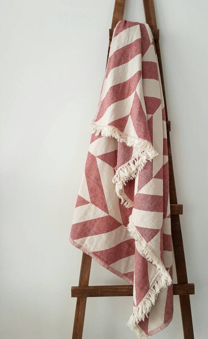 May include: A red and cream geometric patterned throw blanket with fringe trim is draped over a wooden ladder. The blanket features a repeating pattern of angled stripes and triangles.