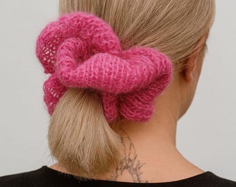 May include: A close-up shot of a person's head and shoulders, with blonde hair pulled back in a low ponytail. A large, fluffy, hot pink scrunchie is wrapped around the ponytail. The person is wearing a black shirt.