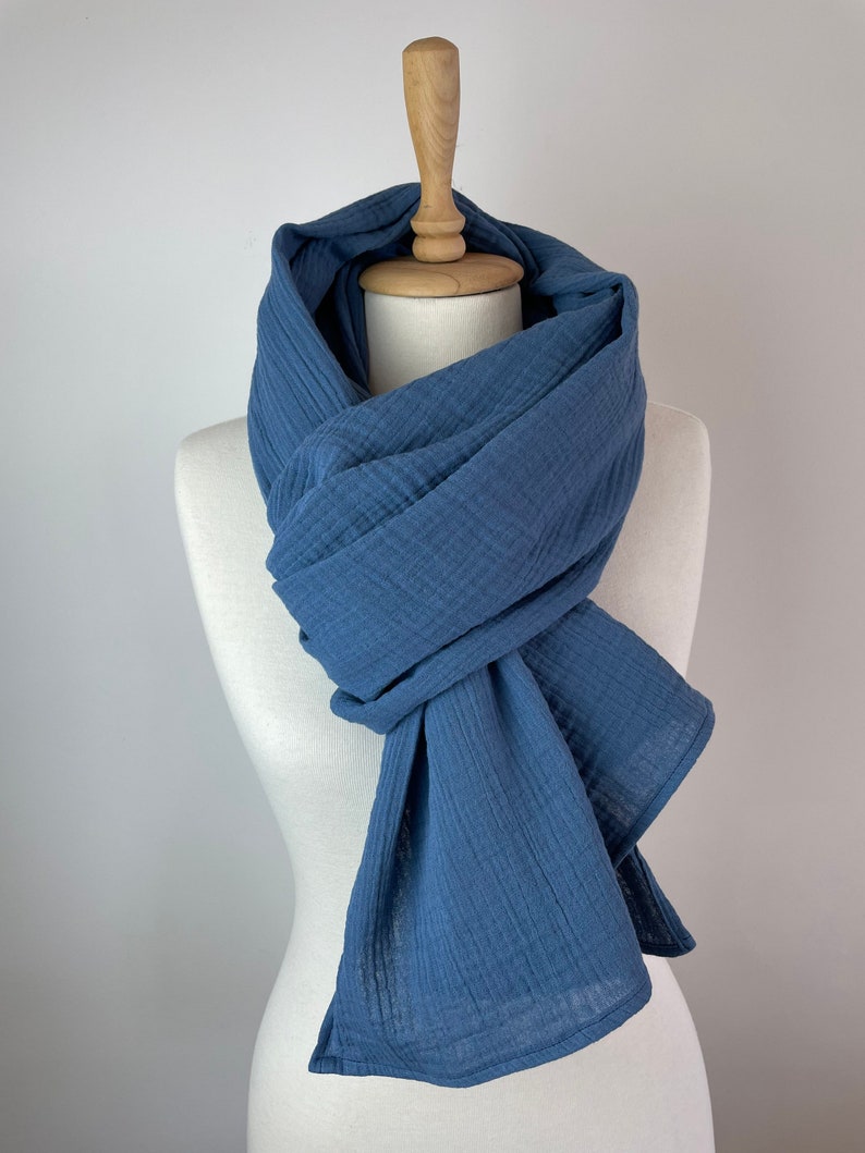 May include: A blue scarf displayed on a mannequin. The scarf, made from textured fabric, is a solid blue. It's wrapped around the mannequin's neck and drapes down. The mannequin is white, with a wooden neck. The scarf's texture is clearly visible.