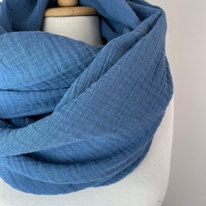 May include: A blue infinity scarf made from a soft, textured material. The scarf is displayed on a mannequin, highlighting its flowing design. The colour is a muted blue, and the fabric looks lightweight and breathable, suitable for cooler weather.