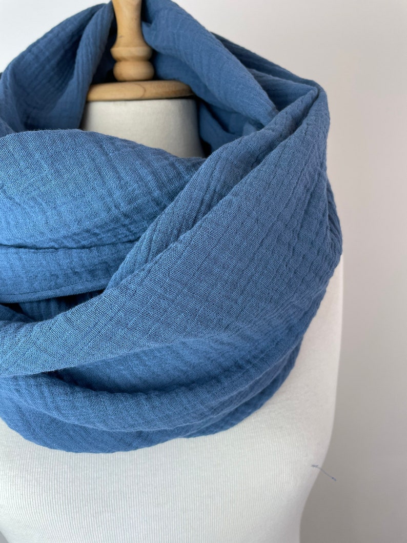 May include: A blue infinity scarf made from a soft, textured material. The scarf is displayed on a mannequin, highlighting its flowing design. The colour is a muted blue, and the fabric looks lightweight and breathable, suitable for cooler weather.
