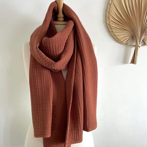 May include: A terracotta-coloured scarf draped over a mannequin. The scarf is made of a textured fabric, possibly gauze or muslin, and has a rectangular shape. The scarf is displayed in a way that suggests it can be worn around the neck or shoulders.