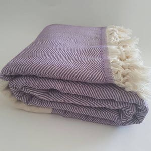 May include: Folded purple and white herringbone-patterned blanket with cream-colored fringe. The blanket is made of a soft material and is folded neatly. The herringbone pattern is a classic design, and the fringe adds a touch of texture.