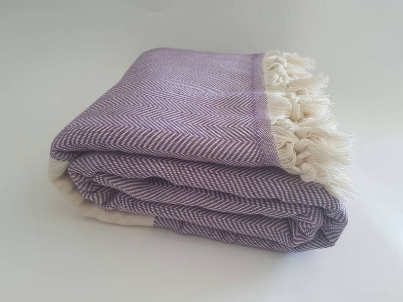 May include: Folded purple and white herringbone-patterned blanket with cream-colored fringe. The blanket is made of a soft material and is folded neatly. The herringbone pattern is a classic design, and the fringe adds a touch of texture.