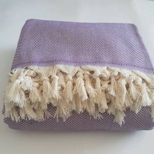 May include: Folded lavender throw blanket with a herringbone pattern and cream-colored fringe. The blanket is made of a soft material and is suitable for use on a bed or sofa. The fringe adds a decorative element.