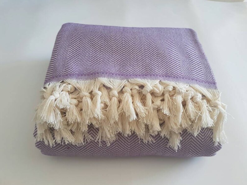 May include: Folded lavender throw blanket with a herringbone pattern and cream-colored fringe. The blanket is made of a soft material and is suitable for use on a bed or sofa. The fringe adds a decorative element.