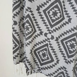 May include: A gray and white patterned textile with a geometric design. The fabric features repeating diamond and zigzag shapes in a dark gray color. The edge has white fringe.
