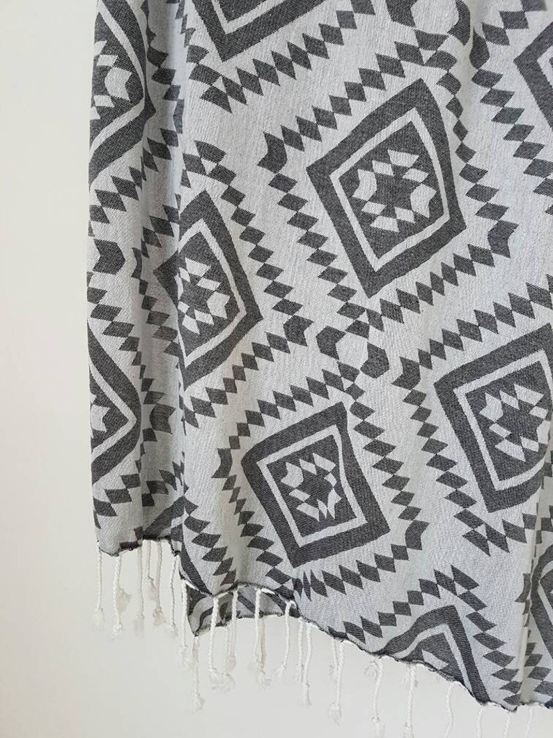 May include: A gray and white patterned textile with a geometric design. The fabric features repeating diamond and zigzag shapes in a dark gray color. The edge has white fringe.