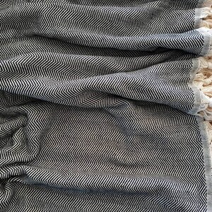 May include: A black and white herringbone patterned blanket with cream-colored fringe. The blanket is folded, showing the texture and design. The fringe is along one edge of the blanket. The blanket is made of a soft material.