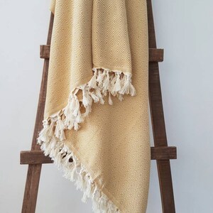 May include: A mustard yellow blanket with a geometric diamond pattern and white fringe. The blanket is draped over a wooden ladder-style rack, showcasing its texture and design. The blanket's edges are finished with a cream-colored fringe.