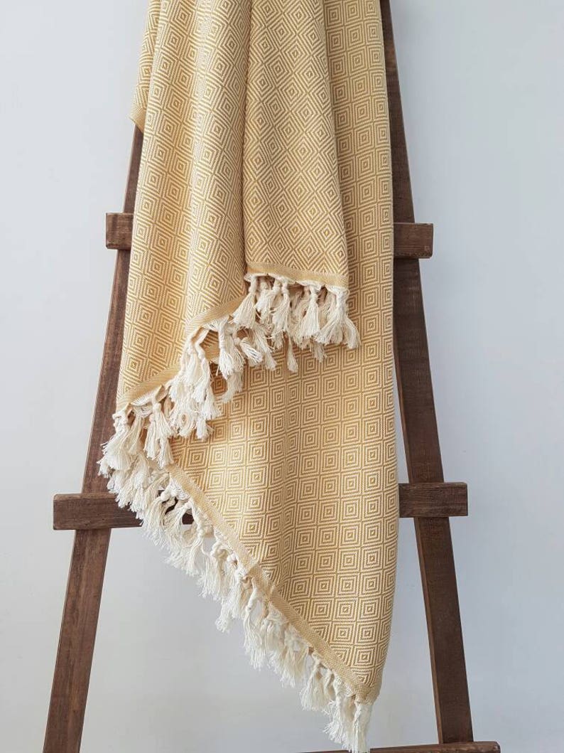 May include: A mustard yellow blanket with a geometric diamond pattern and white fringe. The blanket is draped over a wooden ladder-style rack, showcasing its texture and design. The blanket's edges are finished with a cream-colored fringe.