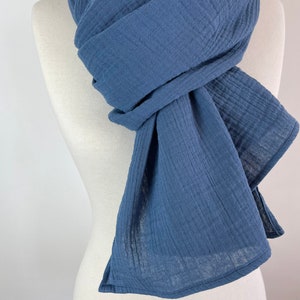 May include: A blue scarf draped over a mannequin. The scarf is made of a soft, textured fabric, with visible vertical lines. The scarf is wrapped and knotted, with the ends hanging down. The color is a muted, medium blue.