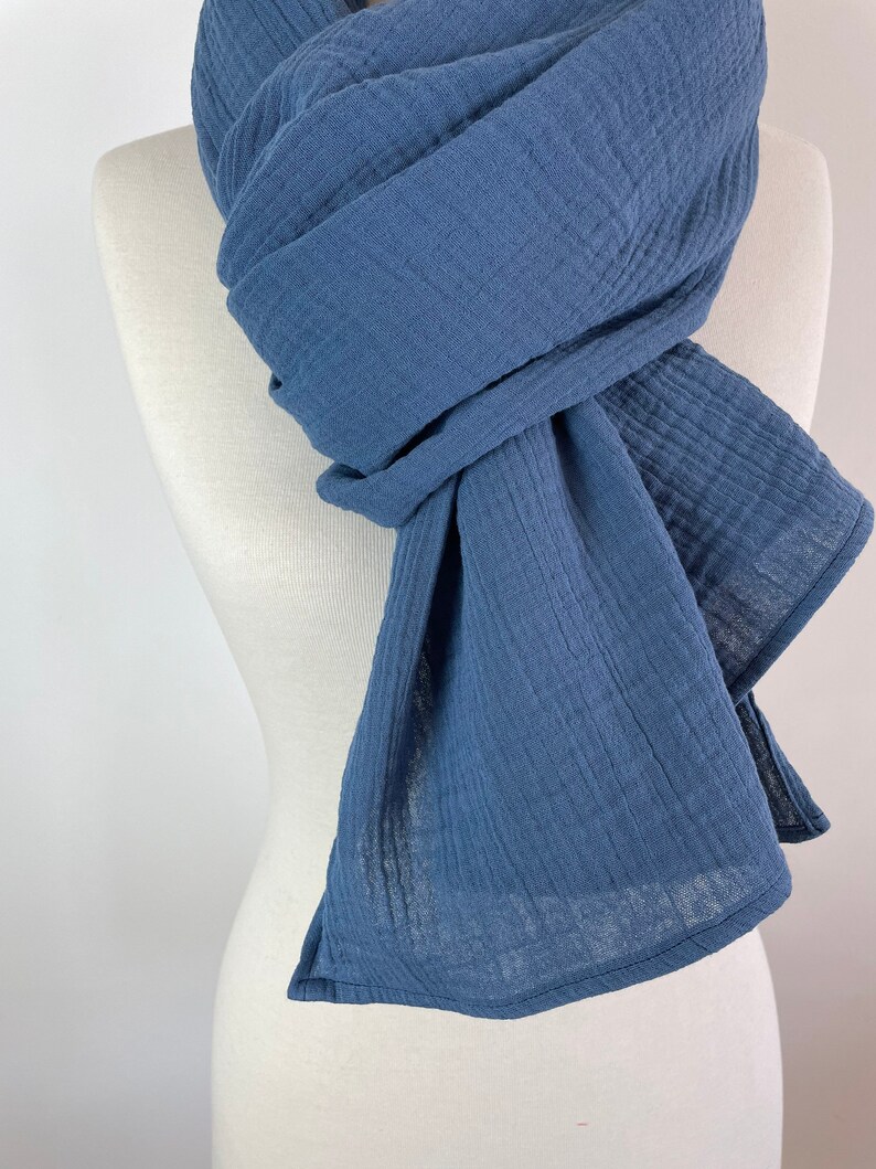 May include: A blue scarf draped over a mannequin. The scarf is made of a soft, textured fabric, with visible vertical lines. The scarf is wrapped and knotted, with the ends hanging down. The color is a muted, medium blue.