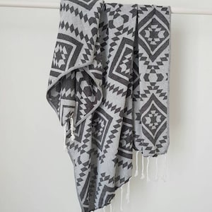 May include: A gray and charcoal geometric patterned towel with white fringe hangs against a white background. The towel features a repeating diamond and triangle design. The towel is made of a soft material.