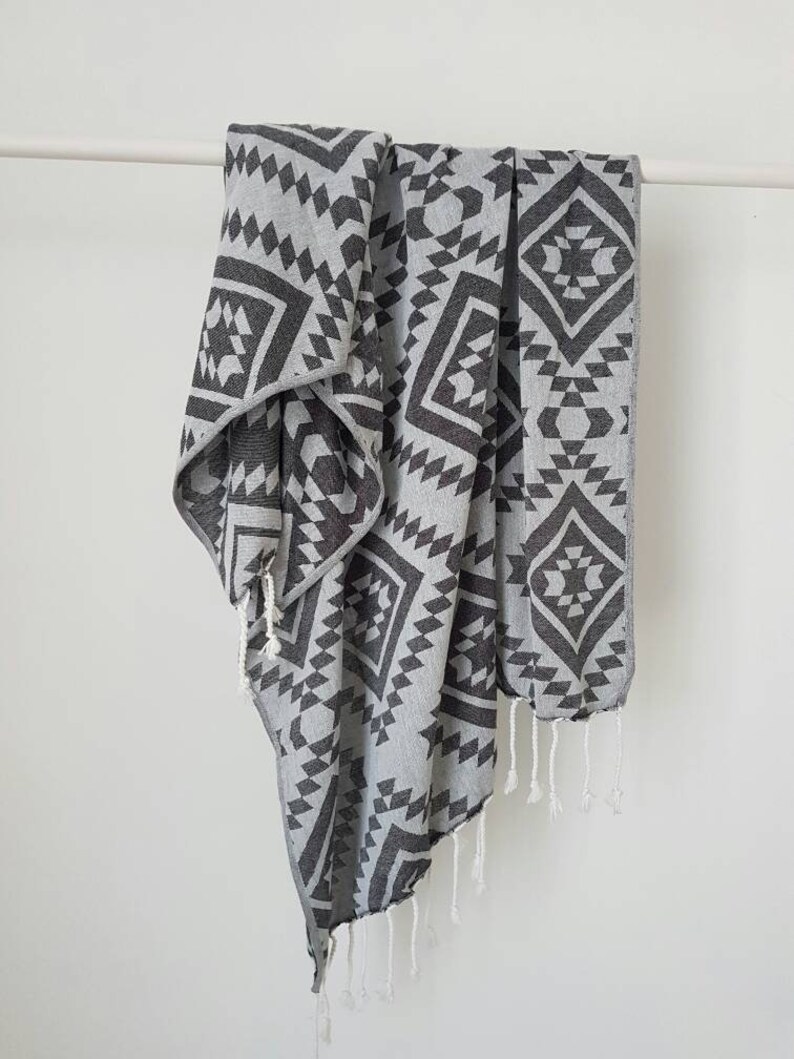 May include: A gray and charcoal geometric patterned towel with white fringe hangs against a white background. The towel features a repeating diamond and triangle design. The towel is made of a soft material.