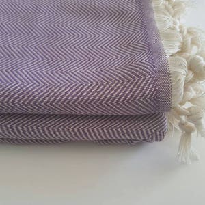 May include: Folded purple and white herringbone-patterned textile with a fringe of white tassels. The fabric is a soft purple with a white zigzag design. The tassels are thick and knotted.