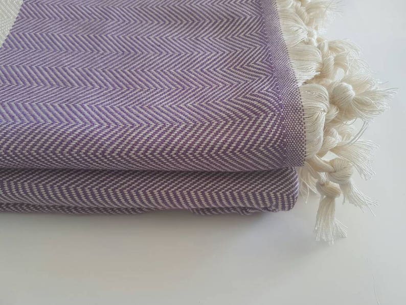 May include: Folded purple and white herringbone-patterned textile with a fringe of white tassels. The fabric is a soft purple with a white zigzag design. The tassels are thick and knotted.