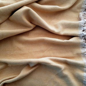 May include: A folded, golden-yellow blanket with a herringbone pattern. The blanket has a fringe of cream-colored tassels along one edge. The fabric is soft and appears to be woven. The blanket is suitable for home decor.